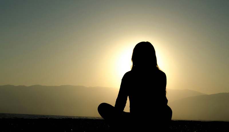 Woman meditating peacefully on a yoga mat near a sunlit window with plants and soft cushions